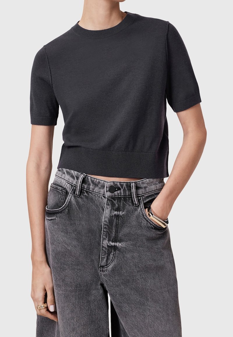 Person som bär en åtsittande svart kortärmad stickad topp och högmidjade urtvättade svarta jeans, med handen i fickan och minimal smyckesprydnad.