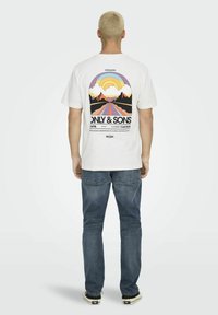 T-shirt bianco in cotone con un design grafico colorato sulla schiena, che raffigura montagne, nuvole e un arcobaleno. Indossato con jeans blu.