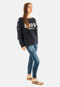 Maglione blu navy con scritte bianche, grandi bottoni testurizzati sulle maniche, abbinato a jeans aderenti e sneakers leggere casual.