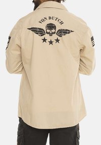 Chemise khaki à boutons avec un graphique de crâne aux ailes et étoiles noires au dos ; comprend des écussons sur les manches avec le drapeau américain.