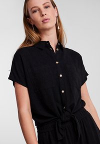 Camicia nera a maniche corte con bottone frontale, due tasche sul petto e un dettaglio annodato in vita. Realizzata in un tessuto texturizzato.