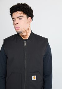 Czarna kamizelka z zamkiem błyskawicznym, przednimi kieszeniami i naszywką z logo Carhartt. Noszona na ciemnej koszuli z długim rękawem, teksturowana tkanina.