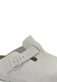Zoccolo in suede grigio con design a T e fibbia regolabile. Presenta una tomaia texture e una soletta marrone per il comfort.