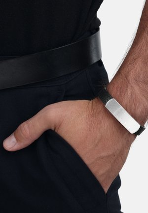 La mano derecha de un hombre con una pulsera de cuero negro y metal descansando en el bolsillo de un pantalón oscuro, lleva un cinturón negro y una camisa negra.