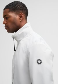 Weiße wasserdichte Jacke mit hohem Kragen, schwarzem Reißverschluss und schwarner Emblem auf dem linken Ärmel. Glatte Textur mit minimalistischem Design.
