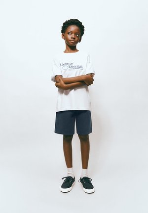 Bambino in piedi con le braccia incrociate, indossa una maglietta bianca "Getaway Essentials", pantaloncini blu navy, calzini bianchi e scarpe da ginnastica nere su uno sfondo semplice.