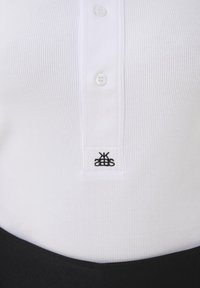 Gros plan sur une chemise blanche côtelée avec trois boutons blancs et un petit logo noir brodé au-dessus de la ceinture noire.