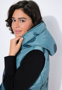 Türkiser Puffer-Weste mit Kapuze, die eine glänzende Oberfläche und dunkle, runde Muster aufweist, über einem schwarzen Langarmoberteil getragen.