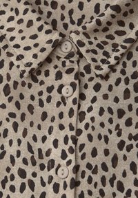 Tessuto beige con una fantasia a stampa leopardata in nero e marrone. Presenta un collo e bottoni chiari lungo la parte anteriore.