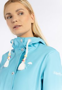 Impermeabilă de culoare albastru deschis, cu guler înalt, accente din mărgele din lemn și un logo de umbrelă albă. Include nasturi cu snapon și șnururi ajustabile.