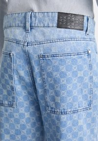 Lyseblå denimjeans med et geometrisk mønster, to baklommer og en svart lærlapp med preget design.