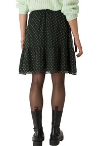 Femme portant une jupe noire à motifs, des collants noirs transparents, des bottes noires épaisses et un pull en tricot vert clair, se tenant avec les mains détendues.