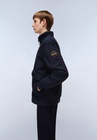 Marineblå jacka med hög krage, sidofickor och en orange cirkulär patch på vänster ärm. Slät textur och minimalistisk design.