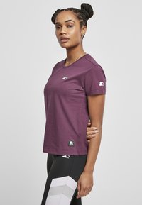 Tricou cu mânecă scurtă, de culoare mov, din bumbac, cu guler rotund. Prezentă un logo pe piept și o etichetă neagră la margine.