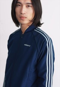 Navy kék zipzáras dzseki, mindkét karon három világoskék csíkkal, az elején, a bal mellrészen hímzett logóval. Sima textúra.