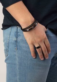 Zwarte leren en zilveren accentarmbanden zijn gestapeld op een pols, gecombineerd met een rechthoekige zwarte ring. De hand rust op lichtblauwe jeans.