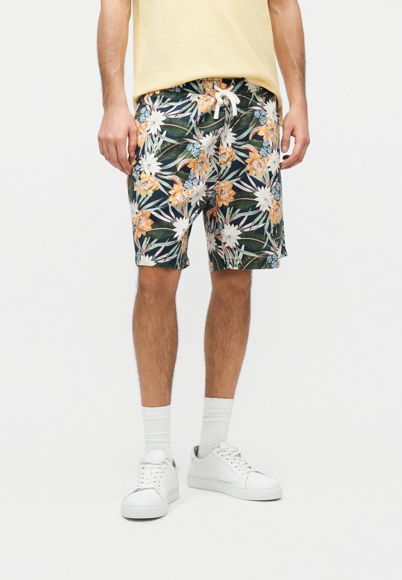 Homme portant un short à motif floral, des baskets blanches, des chaussettes blanches et une chemise jaune, debout devant un fond uni.