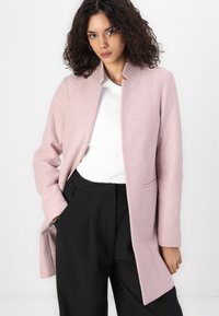 Cappotto di lana rosa chiaro con taglio dritto, senza bottoni e con due tasche laterali, indossato sopra una camicia bianca e pantaloni neri.