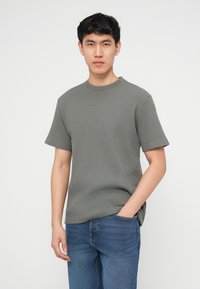 Νεαρός άνδρας με κοντά μαύρα μαλλιά, φορώντας γκρι κοντομάνικο πουκάμισο υφής και μπλε τζιν, στέκεται με το ένα χέρι στην τσέπη.