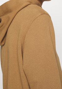 Felpa con cappuccio marrone con una texture morbida, cuciture visibili e cappuccio, mostrata di lato per evidenziare il tessuto e la vestibilità.