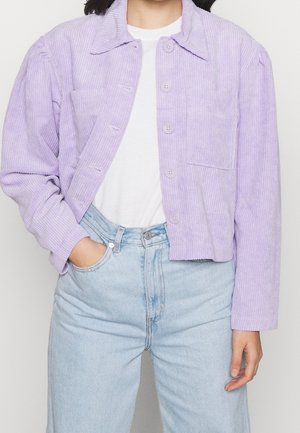 Personne portant une veste en velours côtelé violet clair avec boutons sur une chemise blanche, associée à un jean bleu clair taille haute, avec une main dans la poche.