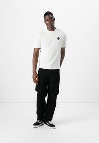 White cotton t-shirt with short sleeves and a small circular logo on the chest, paired with black wide-leg pants and black sneakers.