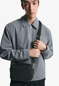 Giacca grigia con zip e tasche laterali, e una borsa a tracolla nera in pelle rettangolare con dettaglio testurizzato e tracolla regolabile.