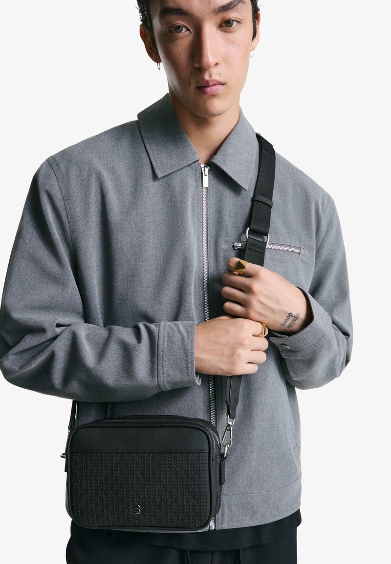 Giacca grigia con zip e tasche laterali, e una borsa a tracolla nera in pelle rettangolare con dettaglio testurizzato e tracolla regolabile.