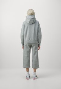 Person med blont hår som bär en grå huvtröja, lösa grå korta byxor, vita strumpor och vita sneakers, står med ryggen vänd mot oss.