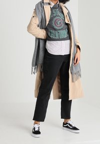 Trinchera beige sobre un sudadera gris y una camisa a rayas, combinada con jeans negros y zapatillas en blanco y negro. Bufanda gris con flecos.