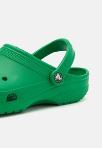 Zoccoli verdi moldati realizzati in materiale leggero, dotati di un cinturino posteriore, aperture laterali perforate e un'accenno di logo circolare.