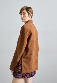 Άτομο με κοντά μαλλιά φοράει ένα καφέ σουέτ μπουφάν πάνω από μια μοτίβα μπορντό φούστα, κοιτάζοντας πίσω σε ένα απλό ανοιχτό γκρι φόντο.