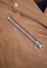 Hellbraune Lederjacke mit diagonalem Reißverschlussfach und silbernem Hardware, glatter Textur und detaillierten Nähten entlang der Nähte.