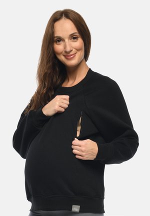Schwangere Frau im schwarzen Sweatshirt demonstriert den Stillzugang, indem sie einen seitlichen Reißverschluss zur Seite zieht und in die Kamera lächelt.