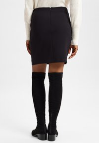 Schwarze, taillierte Röcke aus glattem Material mit einem Reißverschluss hinten, kombiniert mit schwarzen Stiefeln aus Wildleder, die über das Knie reichen und seitliche Reißverschlüsse haben.