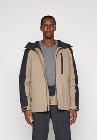 Άνδρας που φοράει μπεζ και μαύρο μπουφάν πάνω από γκρίζο πουκάμισο και σκούρα παντελόνια, στέκεται μπροστά σε απλό ανοιχτό φόντο.