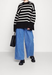 Black and white striped knit sweater with wide sleeves, paired with loose light blue denim trousers and black chunky heeled boots. Black bag.