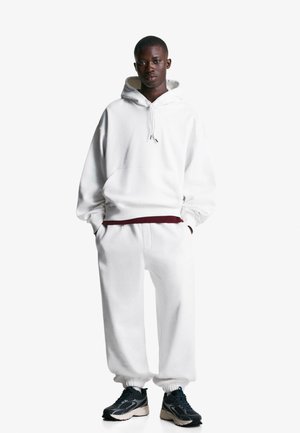 White fleece hoodie with drawstrings, matched with white joggers. Features ribbed cuffs, relaxed fit, and black athletic sneakers.