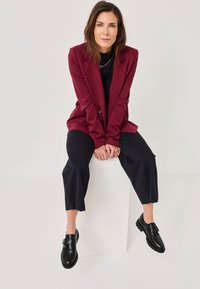 Burgundia bleiser musta kõrge kaelusega sörkide peal, koos laia musta pükstega ja mustade kingadega, millel on rihma detail. Sile kangas, struktureeritud disain.