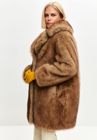Palton din blană sintetică de culoare marron, cu un guler mare și mâneci lungi și ample. Prezentare cu deschidere frontală, purtat cu mănuși galbene, afișat pe un fundal neutru.