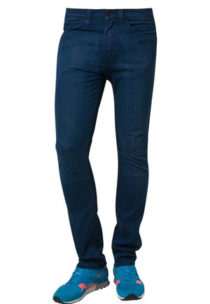 Slim-fit donkerblauwe spijkerbroek gedragen met felblauwe en koraalkleurige sportschoenen, weergegeven van taille tot voeten tegen een witte achtergrond.