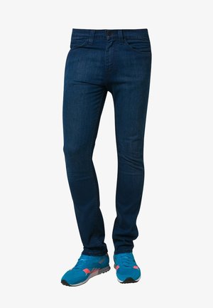 Eng anliegende dunkelblaue Jeans, kombiniert mit leuchtend blauen und korallenfarbenen Sportschuhen, von der Taille bis zu den Füßen auf weißem Hintergrund gezeigt.