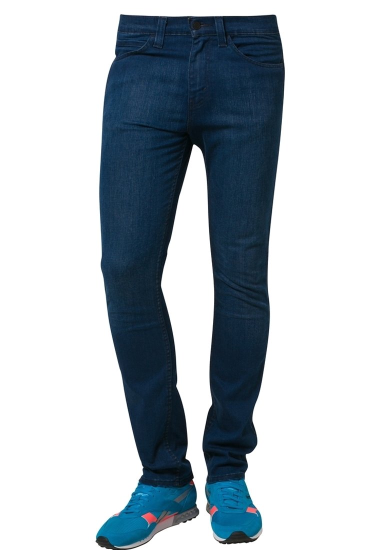 Jean slim bleu foncé porté avec des chaussures de sport bleu vif et corail, vu de la taille aux pieds sur un fond blanc.