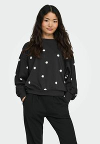 Sieviete ar garām tumšām matiem, ģērbusies melnā mikroplāna džemperī ar baltām margrietiņām un melnās biksēs, stāvot ar rokām kabatās.