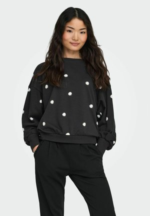 Donna con lunghi capelli scuri, indossa una felpa nera con stampe di margherite bianche e pantaloni neri, in piedi con le mani nelle tasche.