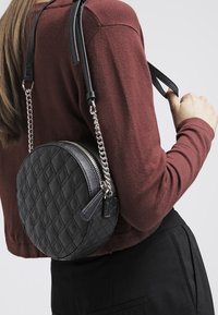 Runde schwarze Handtasche mit gestepptem Muster, Reißverschluss und einer Mischung aus Leder- und Kettenriemen. Über der Schulter getragen, zeigt sie ihre Textur.