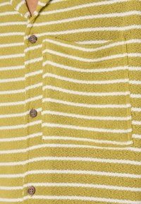 T-shirt en maille rayée de couleur vert olive avec des rayures horizontales blanches. Il possède une poche sur le devant, des boutons et un tissu texturé.