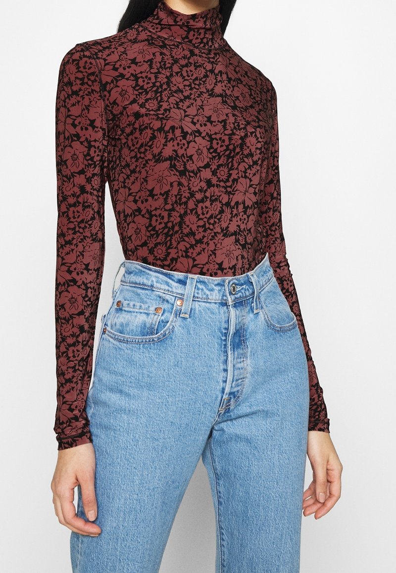 Femme portant un jean taille haute bleu clair et un haut ajusté à manches longues bordeaux avec des motifs floraux noirs et un col cheminée.