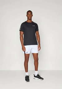 Melns īsām piedurknēm sporta T-krekls ar logo uz krūšu, kombinēts ar baltiem šortiem ar melnu sānu svītru, valkāts kopā ar melniem sporta apaviem.