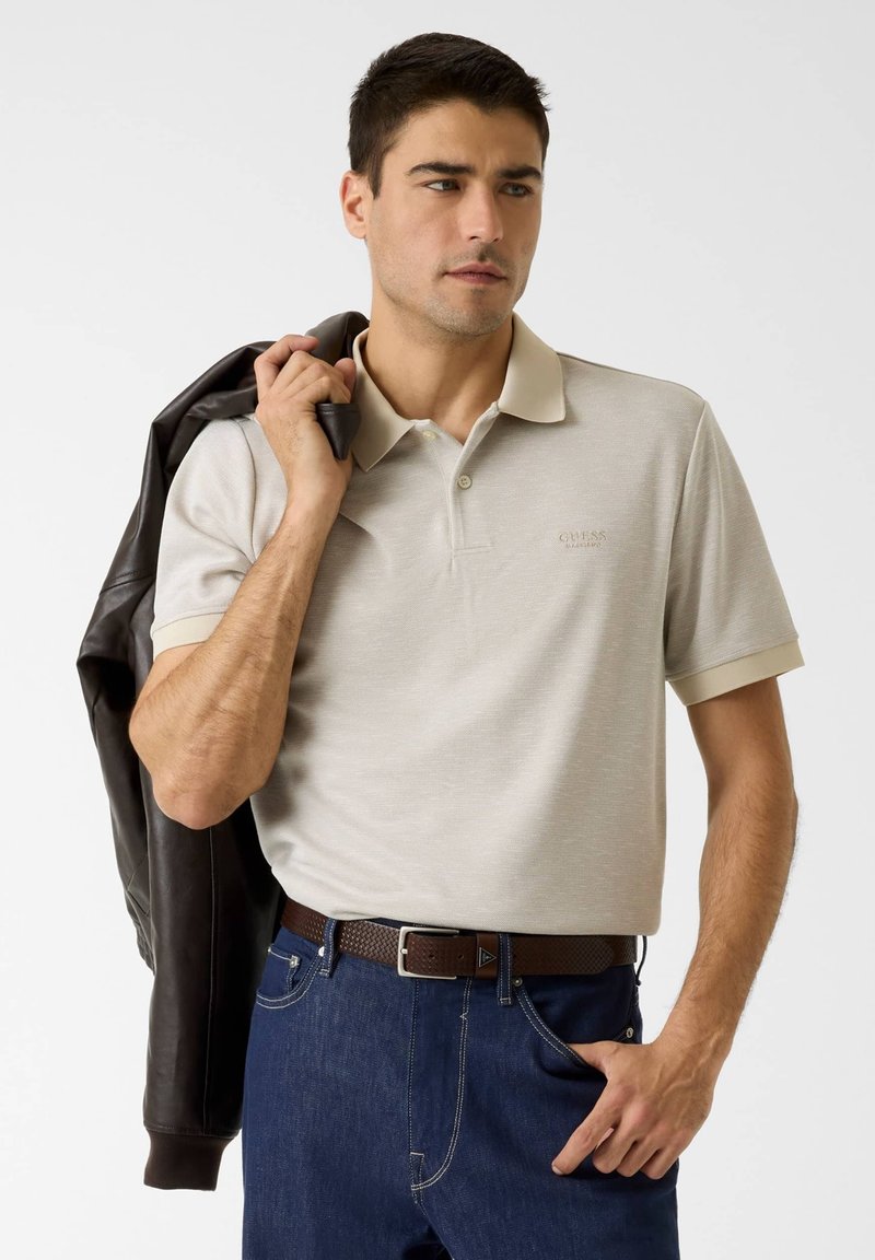 Polo color beige con colletto a contrasto, maniche corte e una texture sottile. Il modello tiene in mano una giacca di pelle scura, con cintura marrone e jeans di denim.